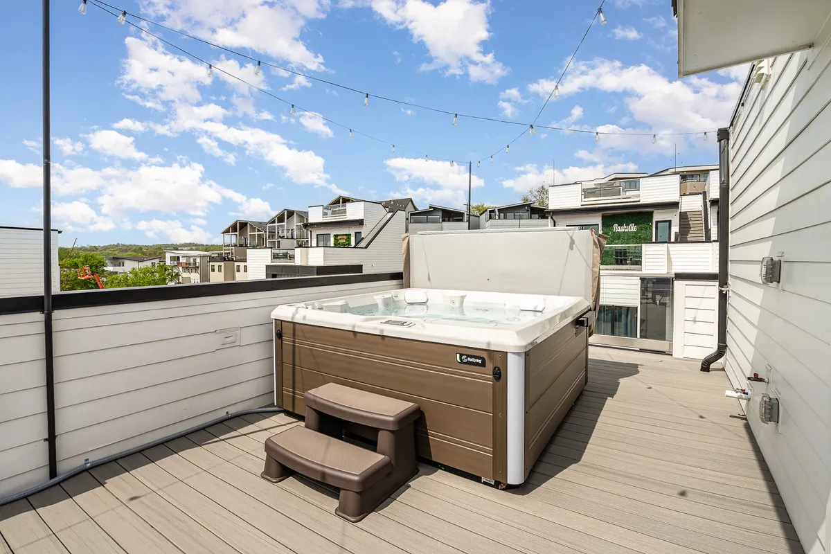 A rooftop hot tub set against the backdrop of surrounding rooftops and string lights, suggesting a relaxing and social evening setting. The view hints at the Nashville skyline, inviting guests to unwind after a night out.