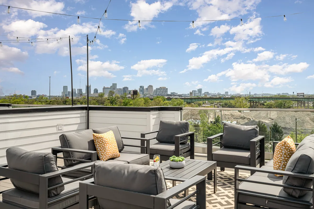 This image captures a contemporary rooftop seating area with decorative cushions, glowing string lights, and sweeping views of the city skyline at dusk. The scene radiates a relaxed, upscale vibe, perfect for gatherings or unwinding while taking in the city lights.