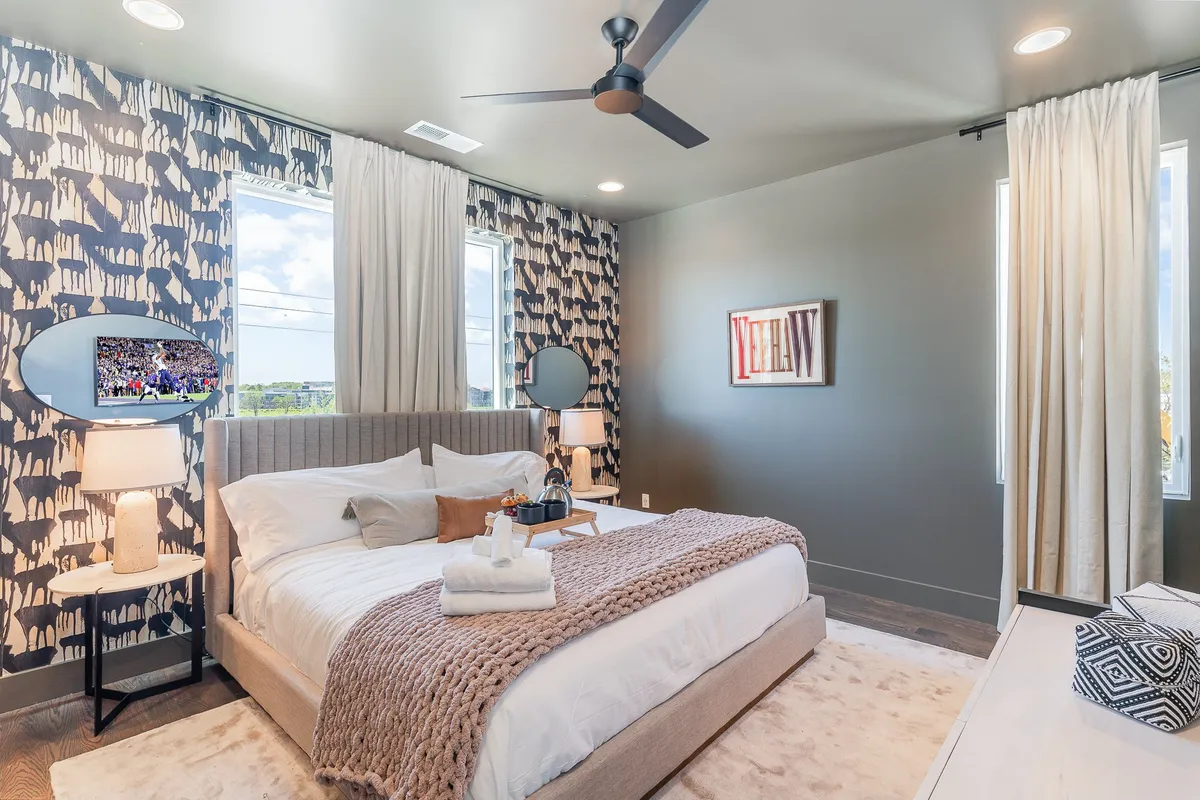 A serene bedroom featuring a plush bed adorned with decorative pillows and a throw blanket. Natural light streams in through the window, illuminating the stylish wall art and soft curtains. A mirrored accent area enhances the sense of space, creating a calming and inviting atmosphere.
