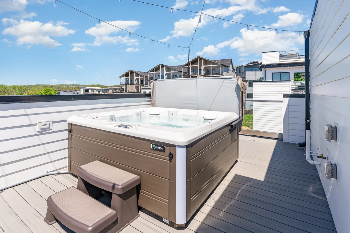 An inviting exterior scene features a bubbling hot tub nestled on an outdoor patio, with warm string lights overhead and steps leading to the deck. The mood is tranquil and intimate, ideal for unwinding or socializing after a busy day.