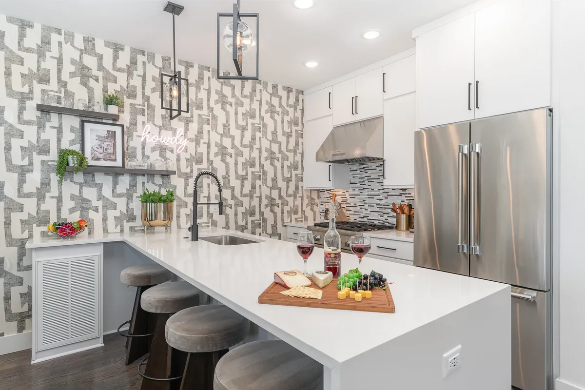 A modern kitchen island set for entertaining, complete with wine and a cheese platter. Sleek stainless steel appliances, stylish textured walls, and hanging light fixtures emphasize a chic and social atmosphere. Bar seating invites guests to gather around the island for casual conversation or shared meals.
