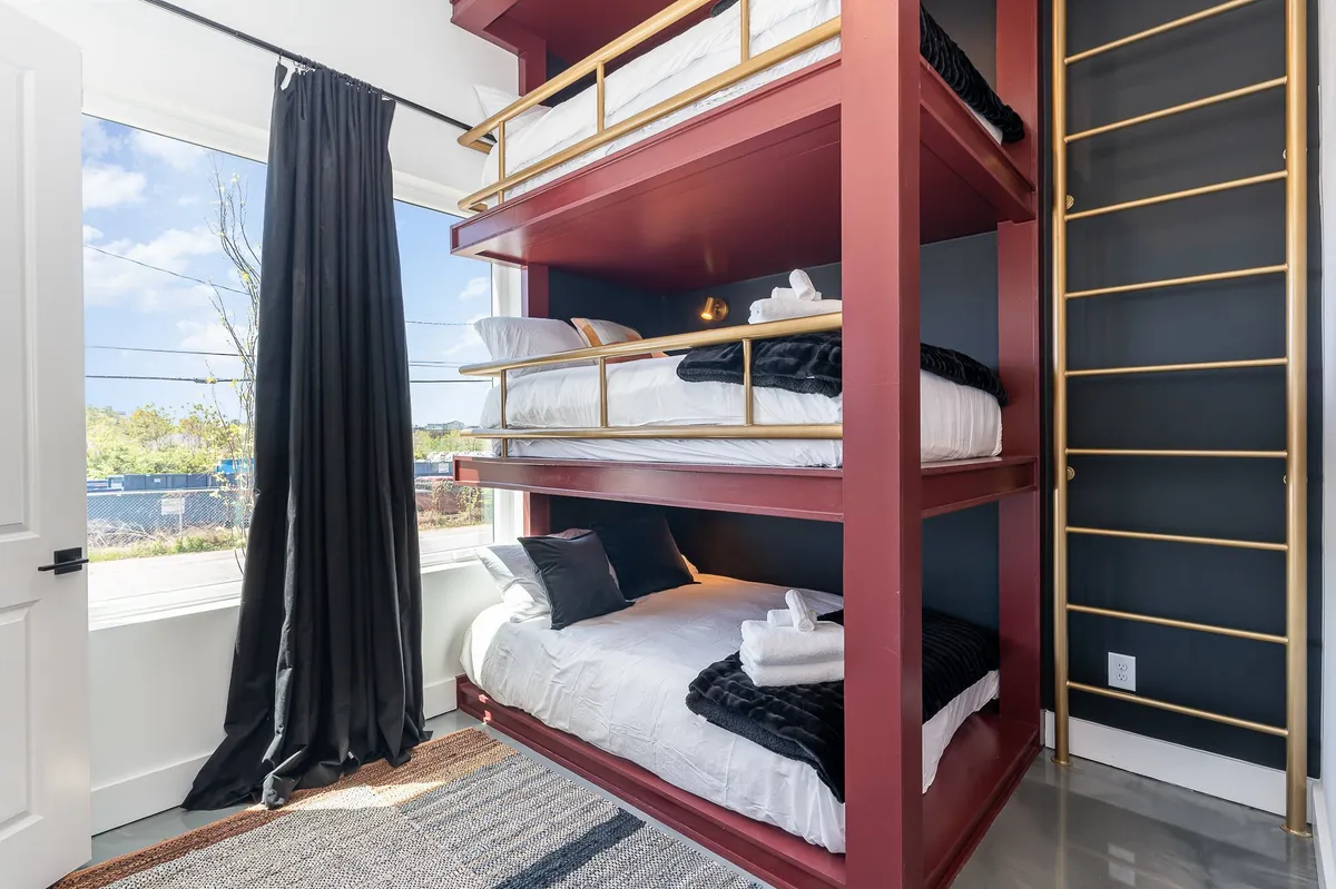 A playful bedroom with sturdy bunk beds positioned by a large window, black curtains framing the morning sunlight. A woven rug adds warmth, creating an ideal space for children or friends to sleep, play, or read together.