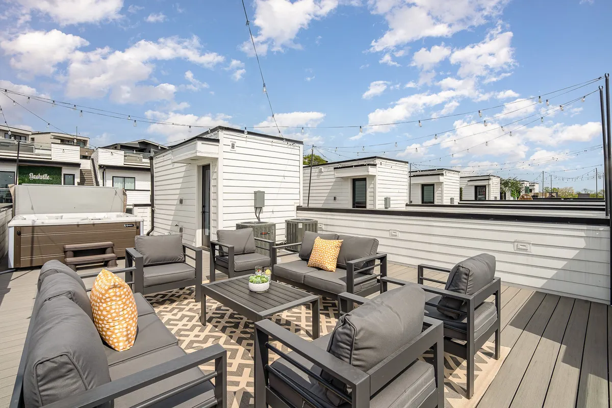 A rooftop scene showcasing a modern outdoor seating area with string lights overhead and a bubbling hot tub, ideal for relaxation and entertaining. The ambiance is cozy and inviting, offering a prime spot for sunset gatherings or starlit soaks.