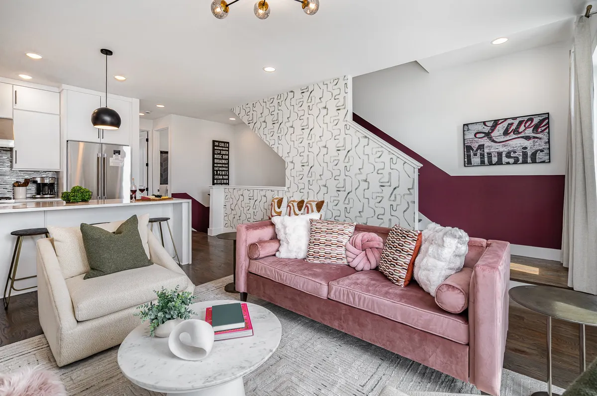 The image showcases a bright, welcoming living area featuring a vibrant pink sofa adorned with decorative pillows, music-inspired artwork on the wall, and a sleek kitchen bar in the background. The space is bathed in natural light, creating a lively atmosphere perfect for gathering and relaxing.