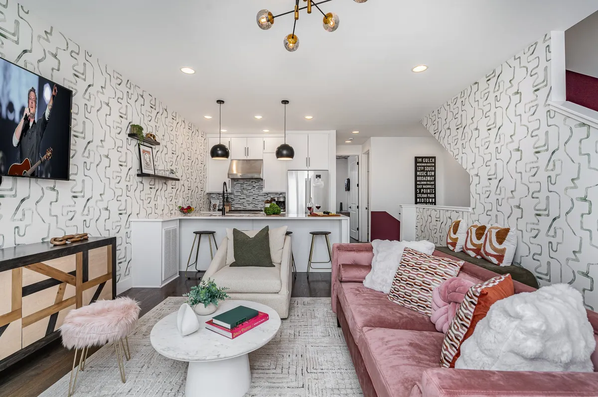 A spacious, stylish living room featuring a sofa with colorful cushions, a white coffee table, a large wall-mounted TV, and a modern open kitchen in the background with visible appliances. The space is well-lit by natural light streaming from large windows, showcasing the seamless flow between living, dining, and kitchen areas.