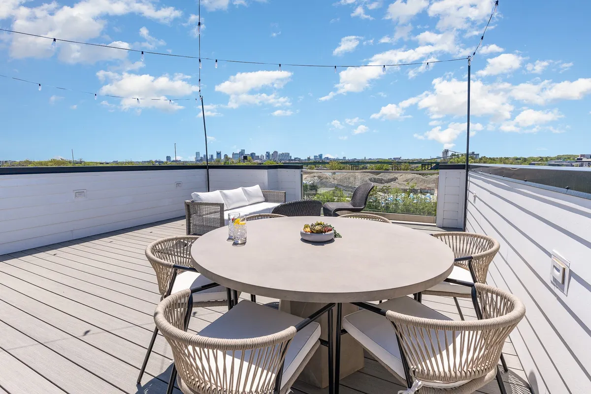 A stylish rooftop dining area featuring a large table set for al fresco meals, city skyline backdrop, and hanging bistro lights across the deck. The mood is casual yet sophisticated, perfect for outdoor dining experiences.