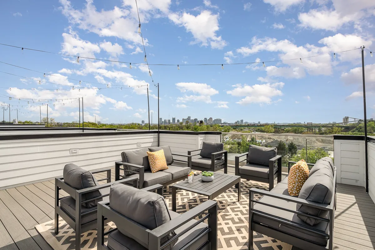 A chic rooftop outdoor seating area is arranged with comfortable chairs, decorative pillows, and string lights. The city skyline is visible in the background, providing an urban yet inviting atmosphere for social gatherings or evening relaxation.